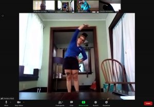 Video chat screen with one large main screen. instructor demonstrating stretch with her hand over her head, other hand on hip and leaning to stretch side. Others in smaller screen are following her instructions.